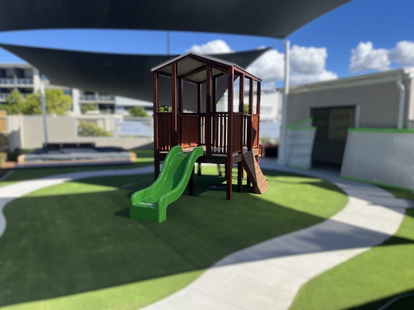 Timber fort with a green slide on synthetic turf with curved concrete paths and shade sails overhead in Birtinya