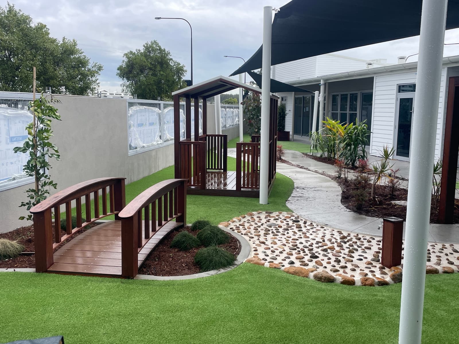 Arched timber footbridge over a planted garden bed beside a pebble creek bed, a small timber cubby and synthetic turf in Birtinya