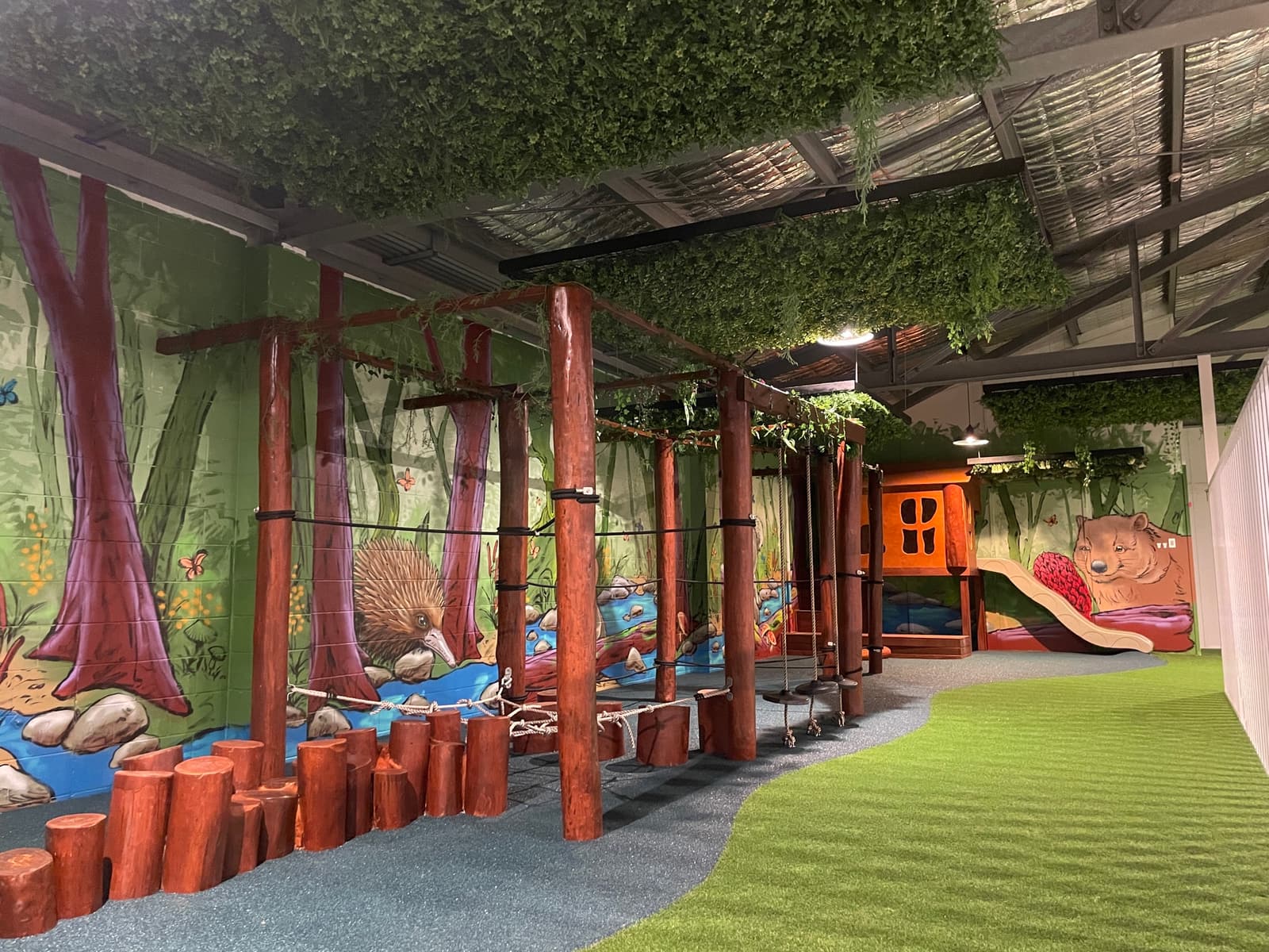 Indoor play space in Byron Bay with a rainforest mural, a timber post and rope obstacle course, log steppers, synthetic turf and grey soft surfacing, and a cubby with a slide at the far end