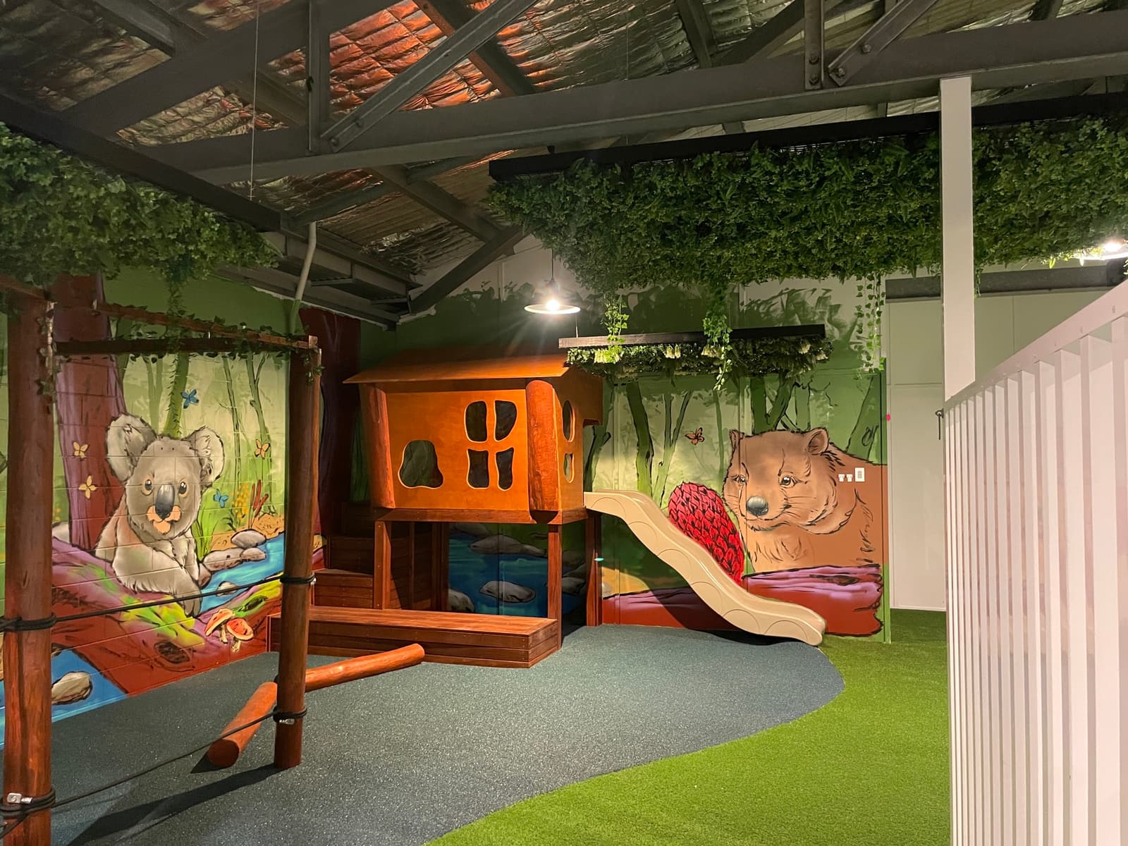 Indoor timber cubby with a cream slide in front of a painted native-animal mural, with artificial foliage overhead, in Byron Bay