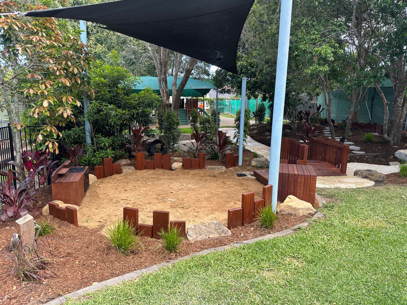 Sandpit edged with a timber log palisade and sandstone boulders under a black shade sail, with garden planting and a small timber bridge, in Carindale