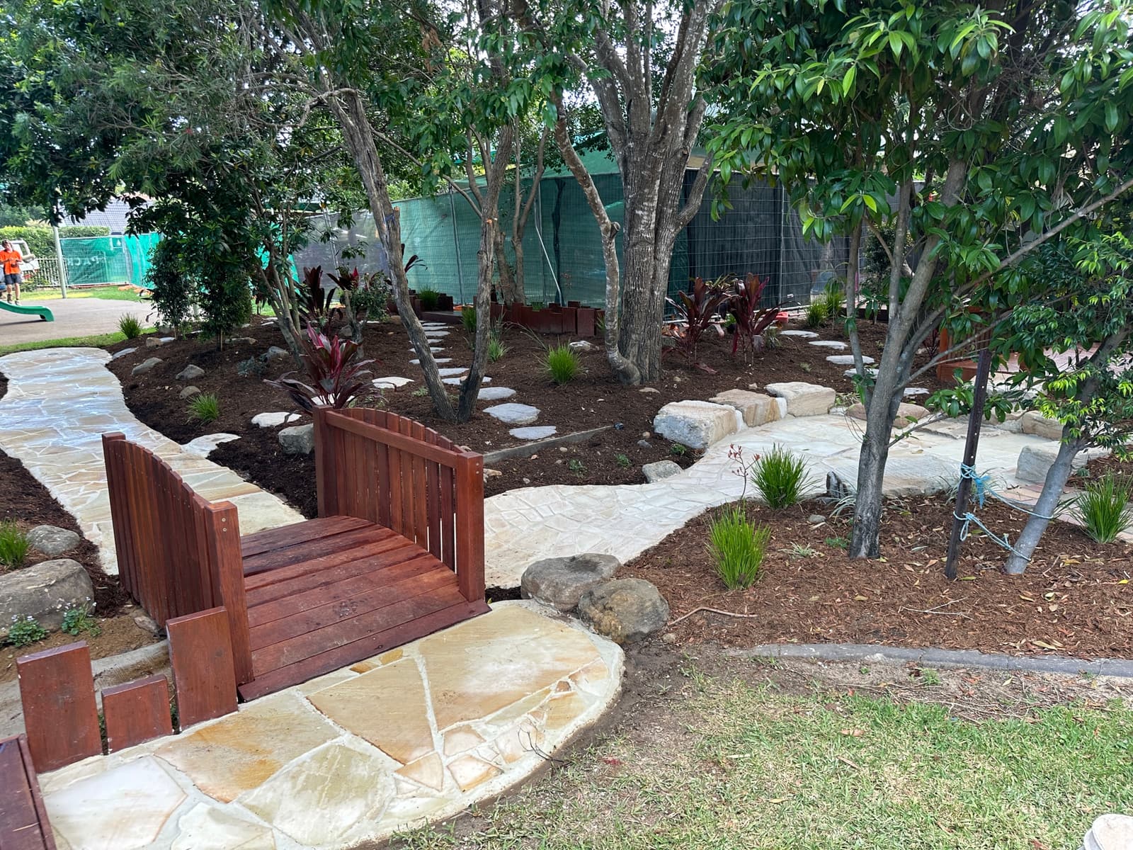 Arched timber bridge on a sandstone crazy-paved path winding through mulched garden beds under established trees in Carindale