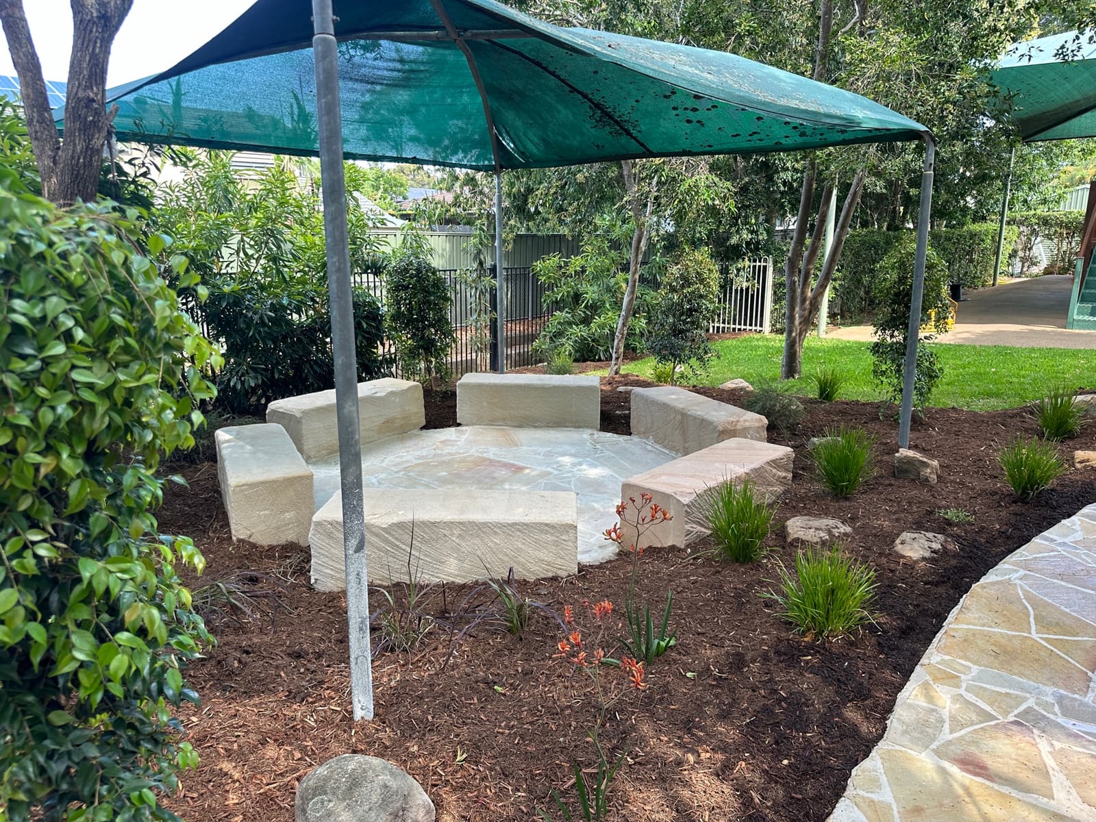 Circle of sandstone block seats on crazy paving in a mulched garden bed beneath a green shade structure in Carindale