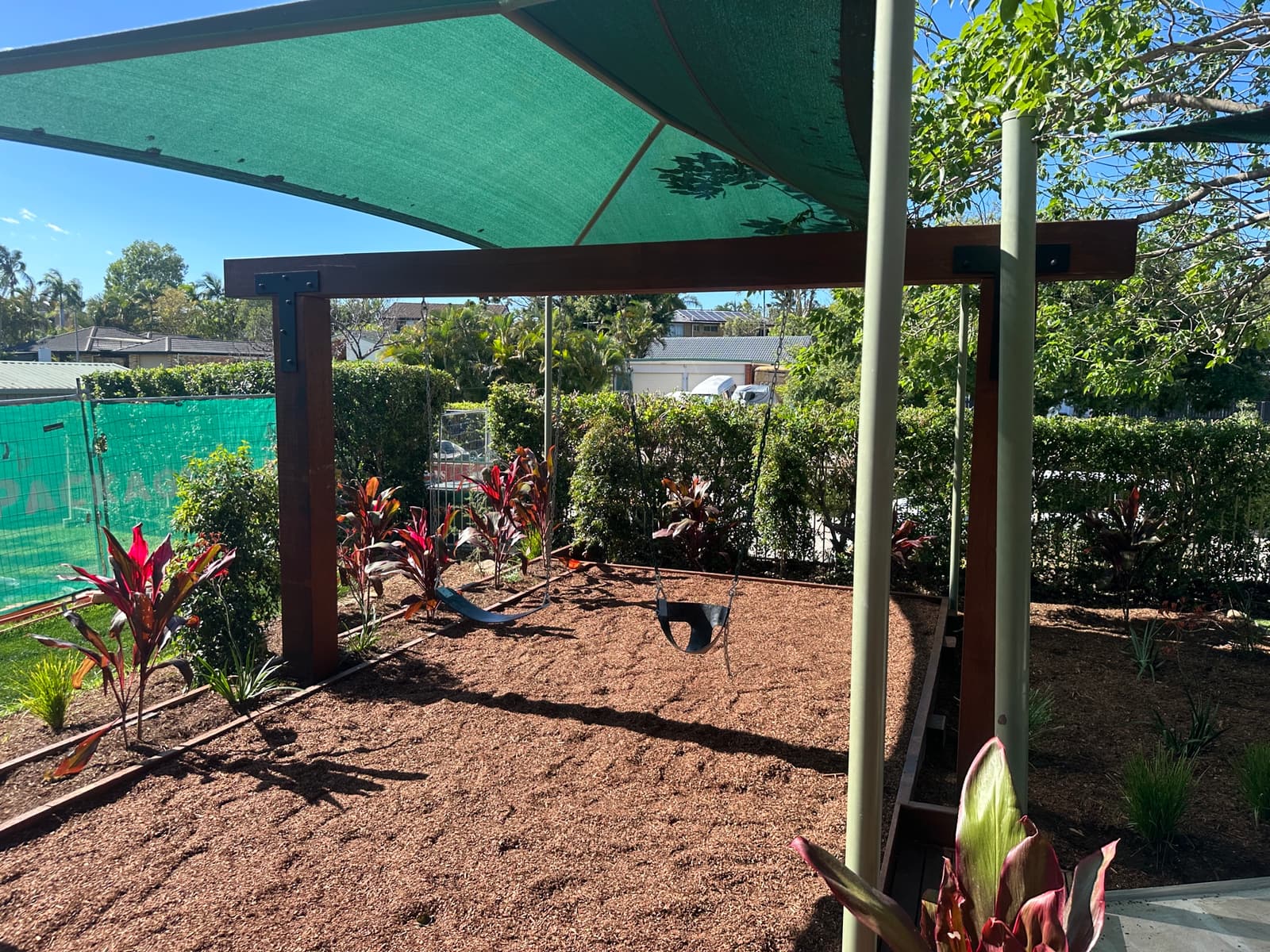 Timber-framed swing set over bark mulch beneath a green shade sail, bordered by planted garden beds, in Carindale