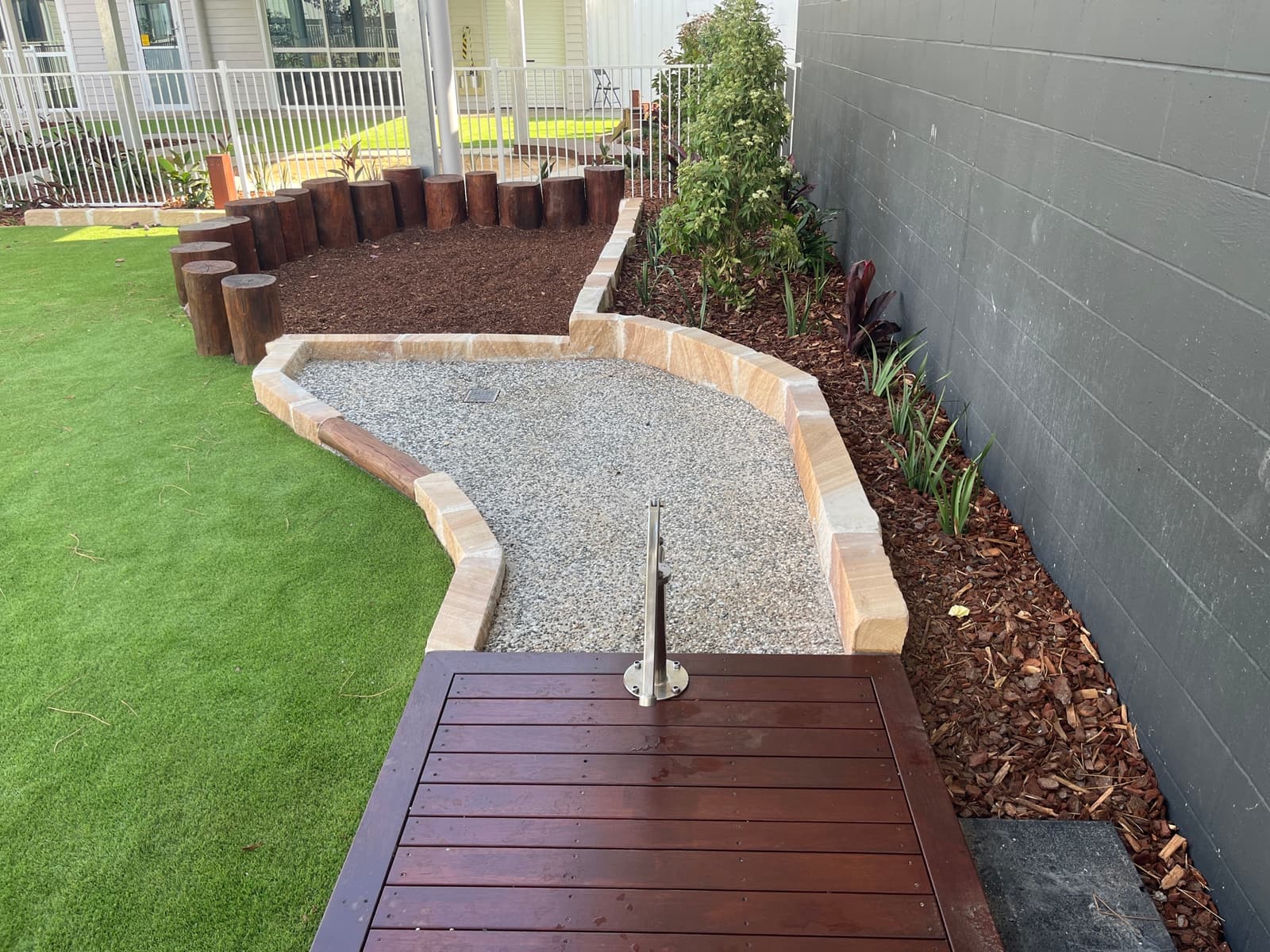 Timber deck leading to a gravel creek bed edged with sandstone blocks, with a log palisade and mulched garden beside synthetic turf, in Cooranbong