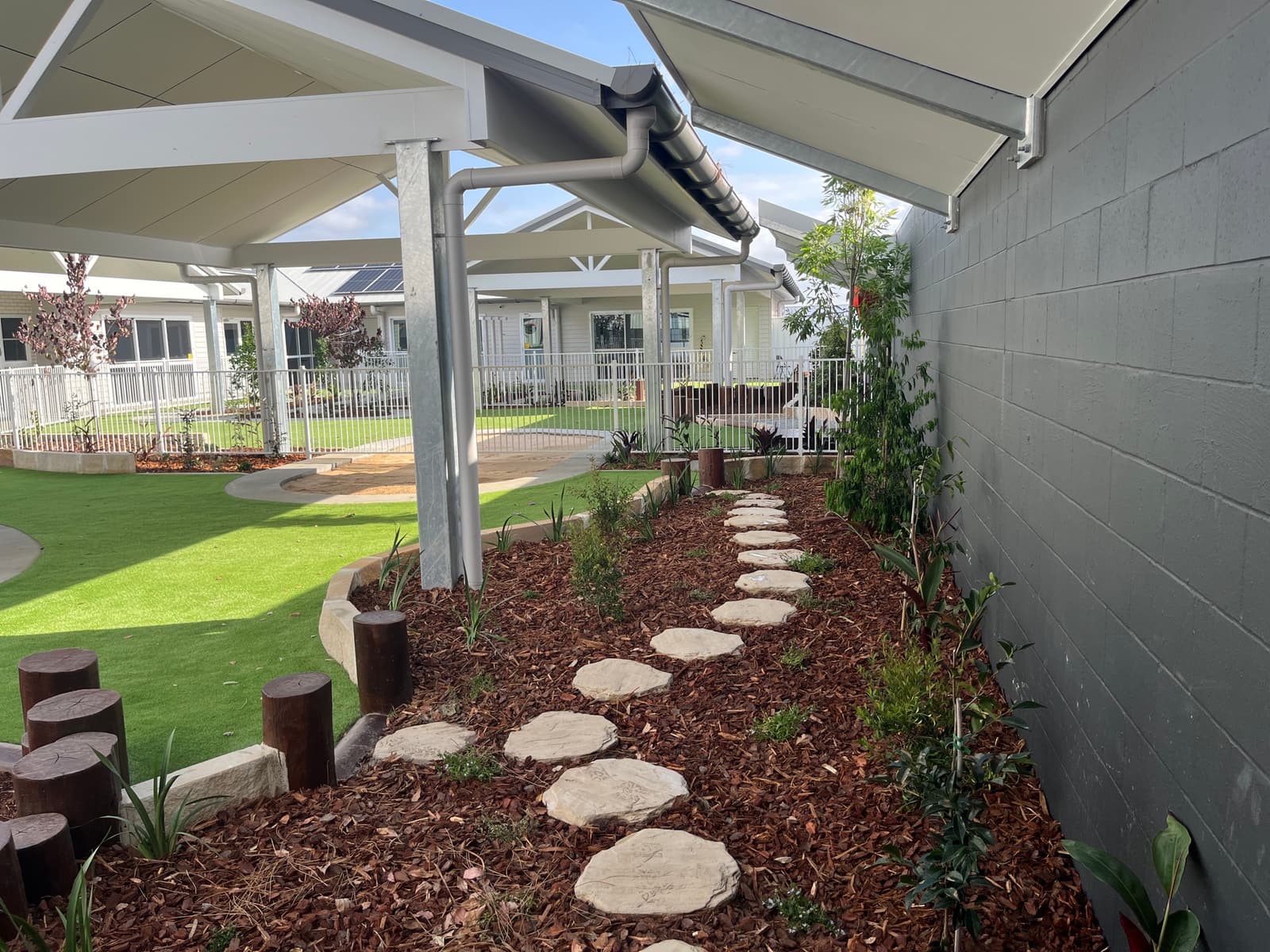 Stepping-stone path through a mulched garden bed with young plants along a wall, with log steppers and synthetic turf beside it, in Cooranbong