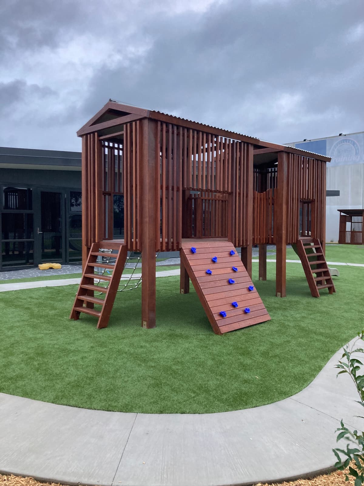 Twin-tower timber fort with slatted walls, a climbing wall with blue holds and two ladders, on synthetic turf beside a concrete path, in Cranbourne