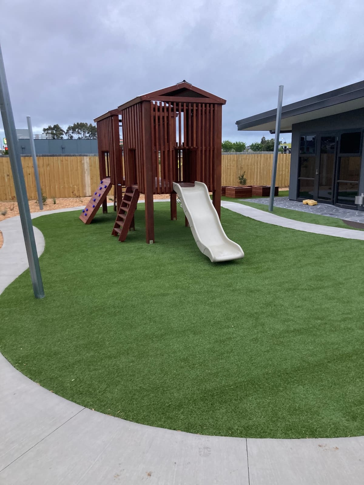 Timber fort with a cream slide, ladder and climbing ramp on synthetic turf ringed by a concrete path in Cranbourne