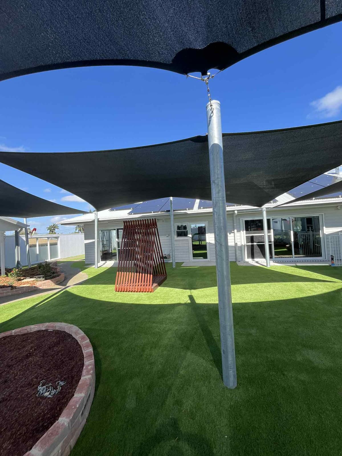 Steel shade sail post and overlapping sails above synthetic turf, with a slatted timber A-frame play structure and a brick-edged garden bed in East Mackay