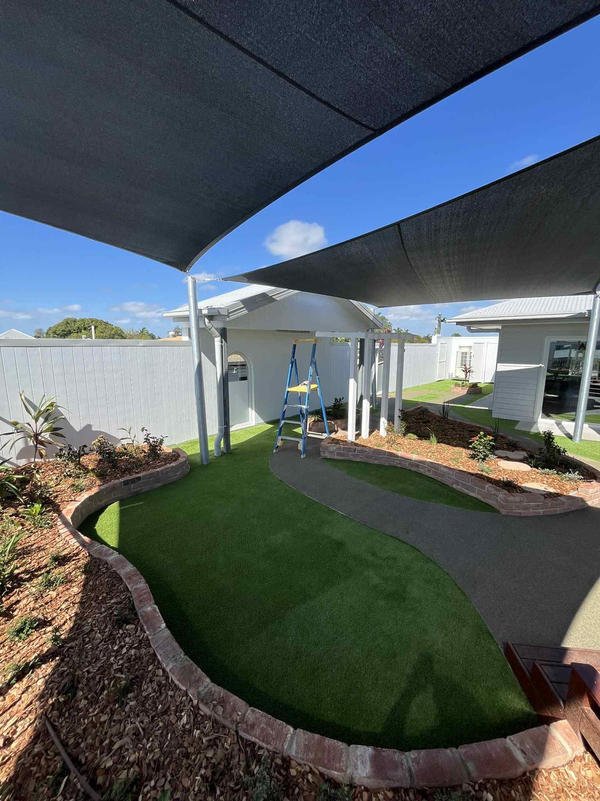 Curving brick garden edging around synthetic turf and a winding dark path under shade sails in East Mackay