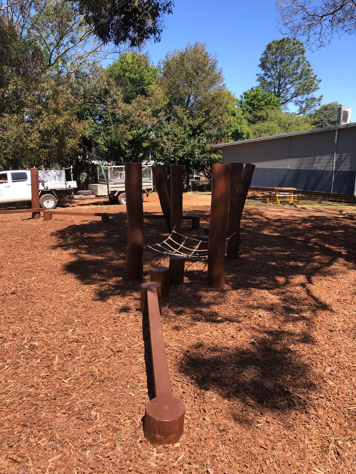 Rope climbing net strung between leaning timber posts, with a low balance beam and log steppers on bark mulch, in Highfields