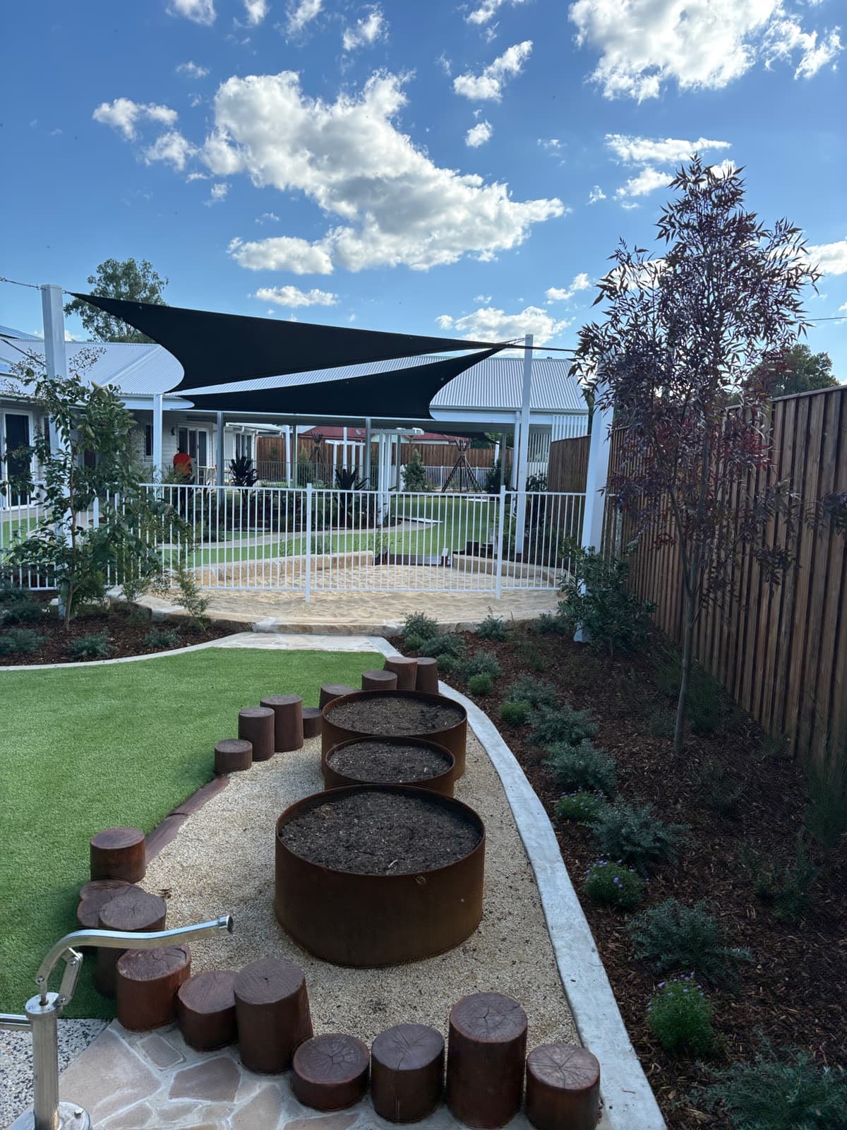 Gravel play path with round steel planters, timber log steppers and a hand water pump beside synthetic turf and garden beds, with shade sails beyond, in Kurri Kurri