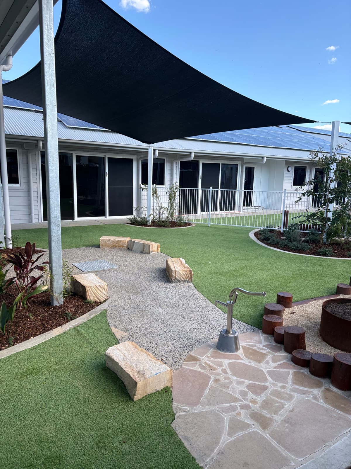 Sandstone crazy paving with a hand water pump, sandstone block seats, a gravel path and synthetic turf under a black shade sail in Kurri Kurri