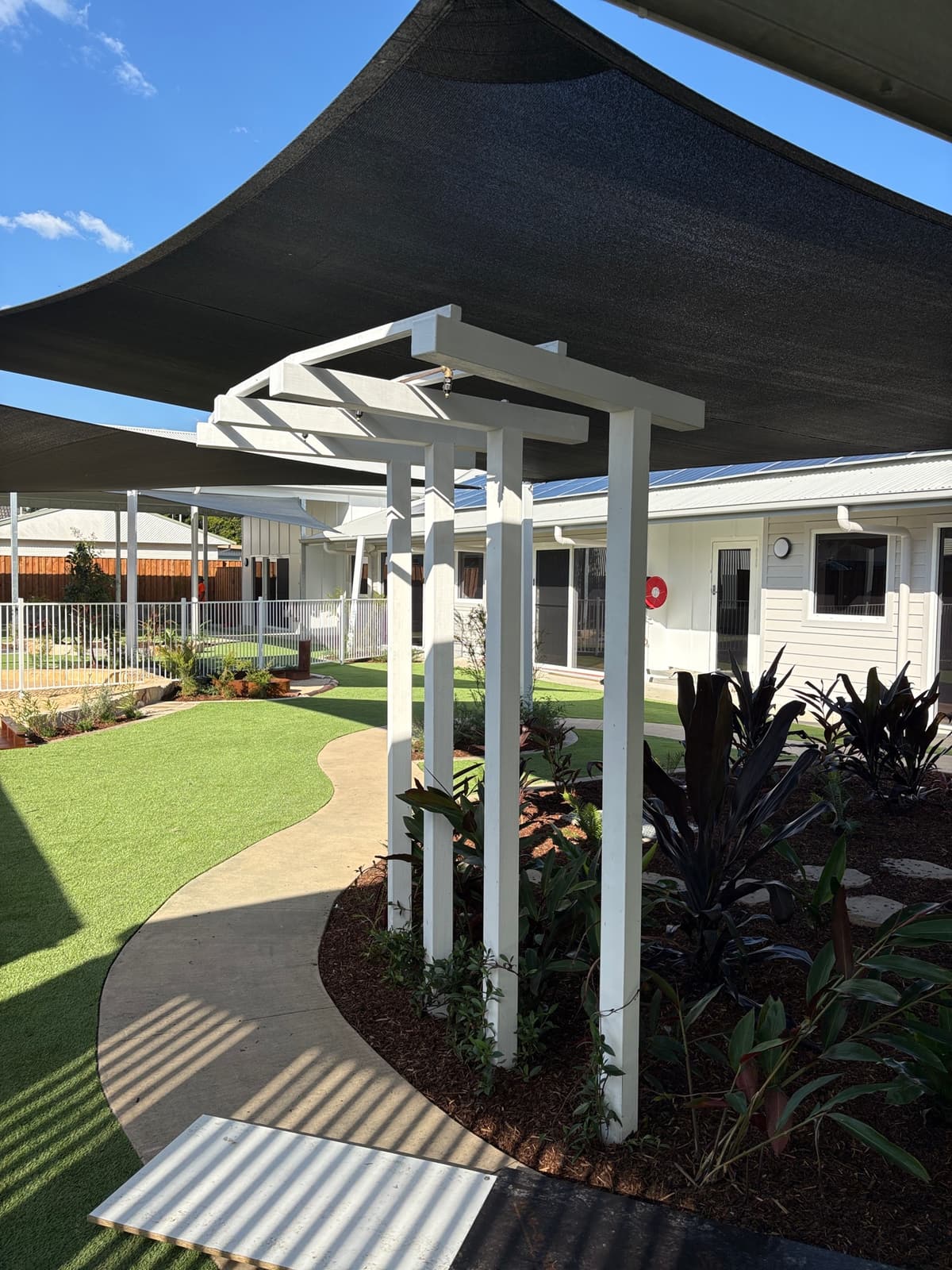 White timber arbour over a curved concrete path through synthetic turf and garden planting, under a shade sail, in Kurri Kurri