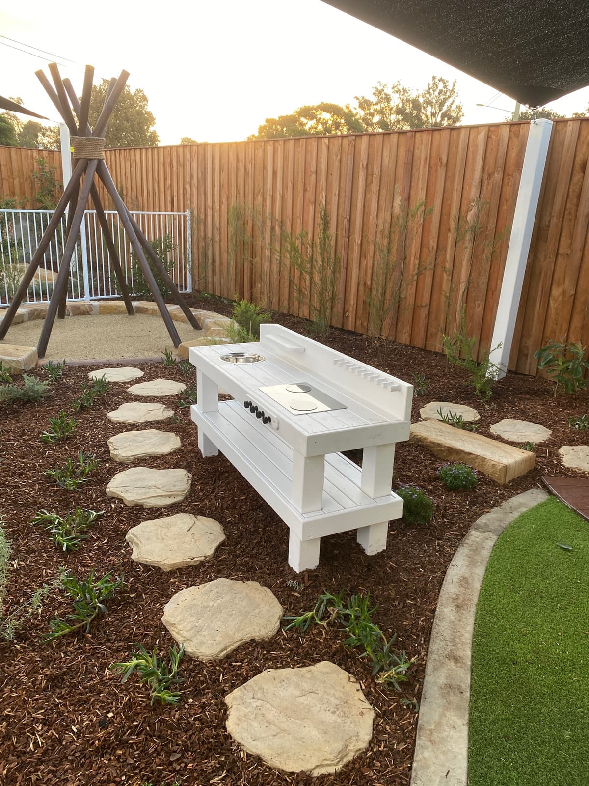 White timber mud kitchen on sandstone stepping stones in a mulched garden bed, with a timber pole teepee behind, in Kurri Kurri