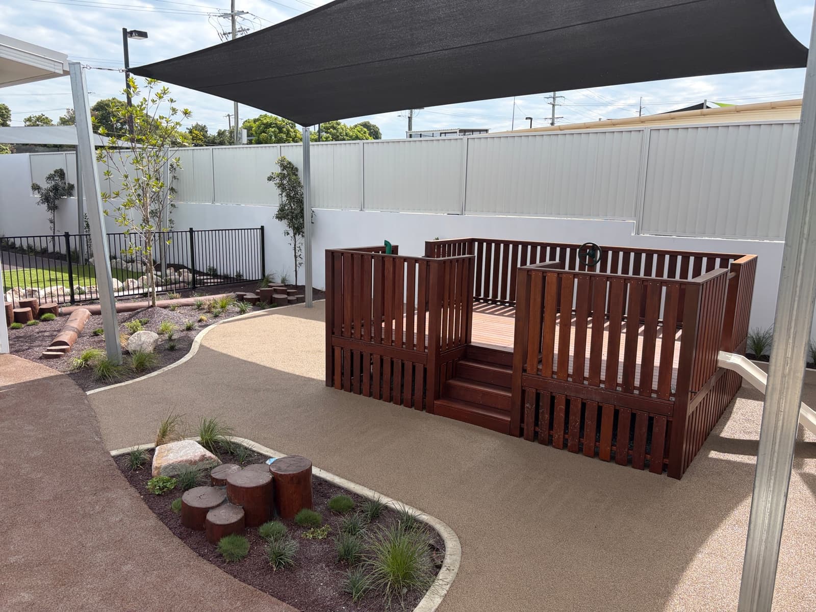 Raised timber deck with picket balustrades and steps on seamless tan play surfacing, with log steppers in a garden bed and a shade sail overhead in Strathpine