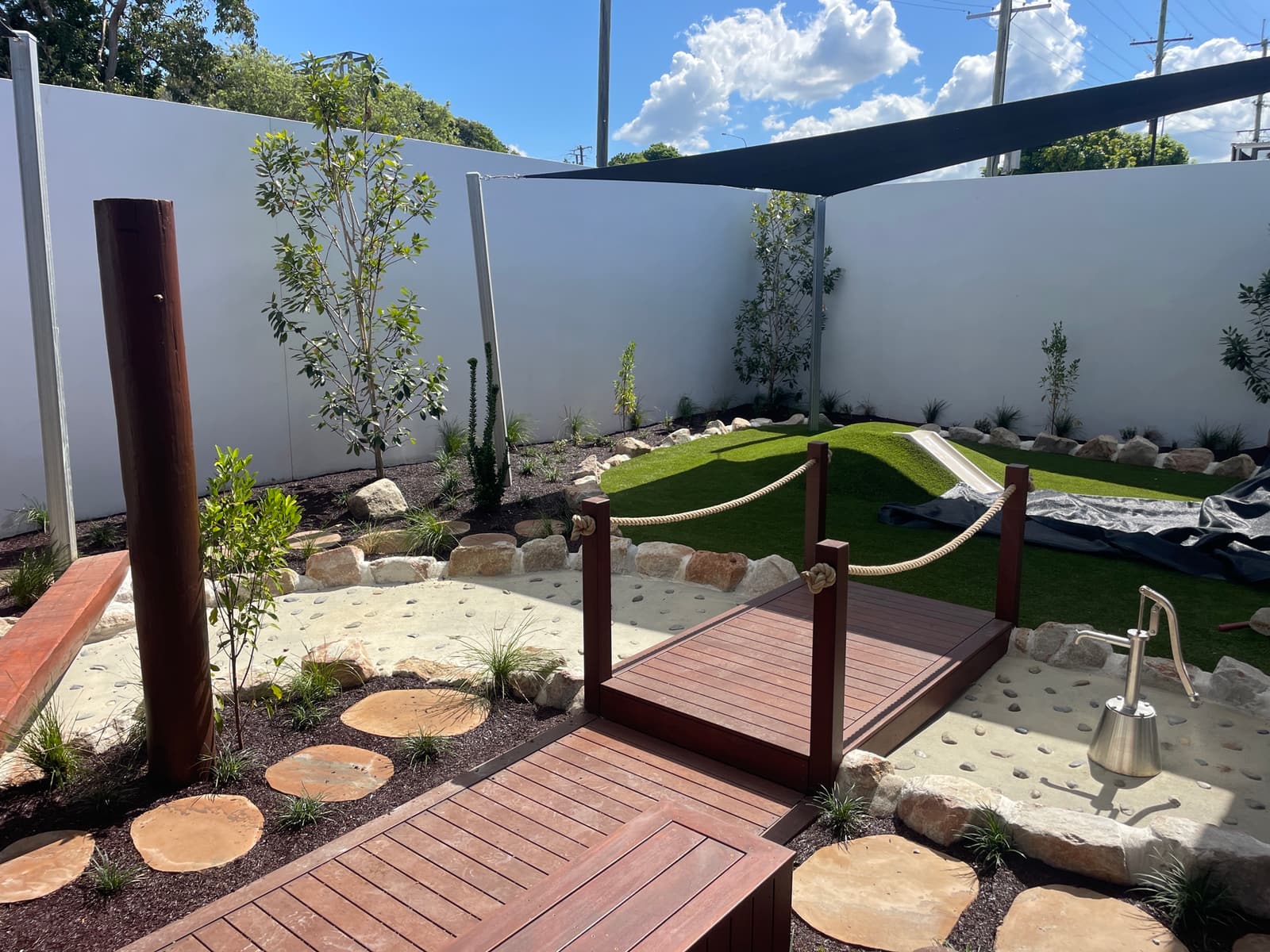 Timber boardwalk bridge with rope handrails crossing a sand and pebble creek bed with a hand water pump, stepping stones and a turf mound under a shade sail in Strathpine