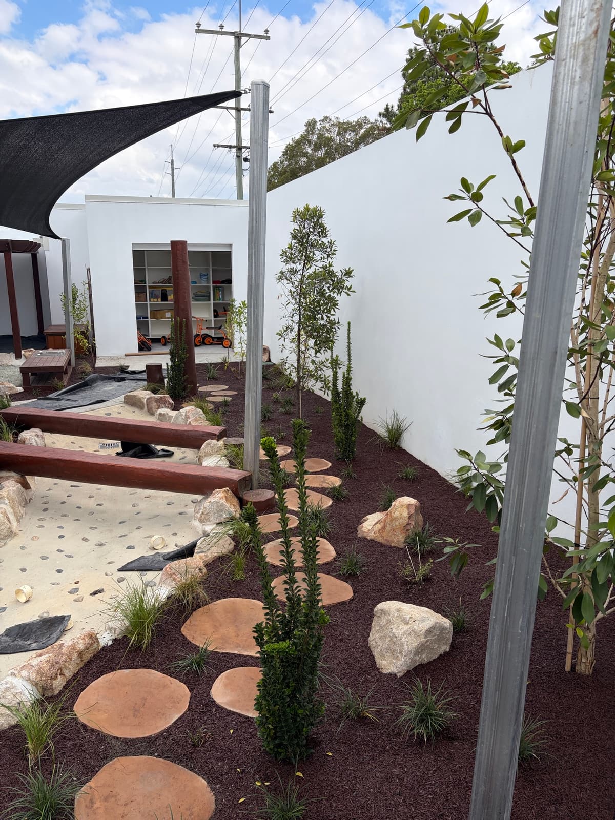 Path of round stepping stones through mulch and young trees beside a sand play area with timber balance beams in Strathpine