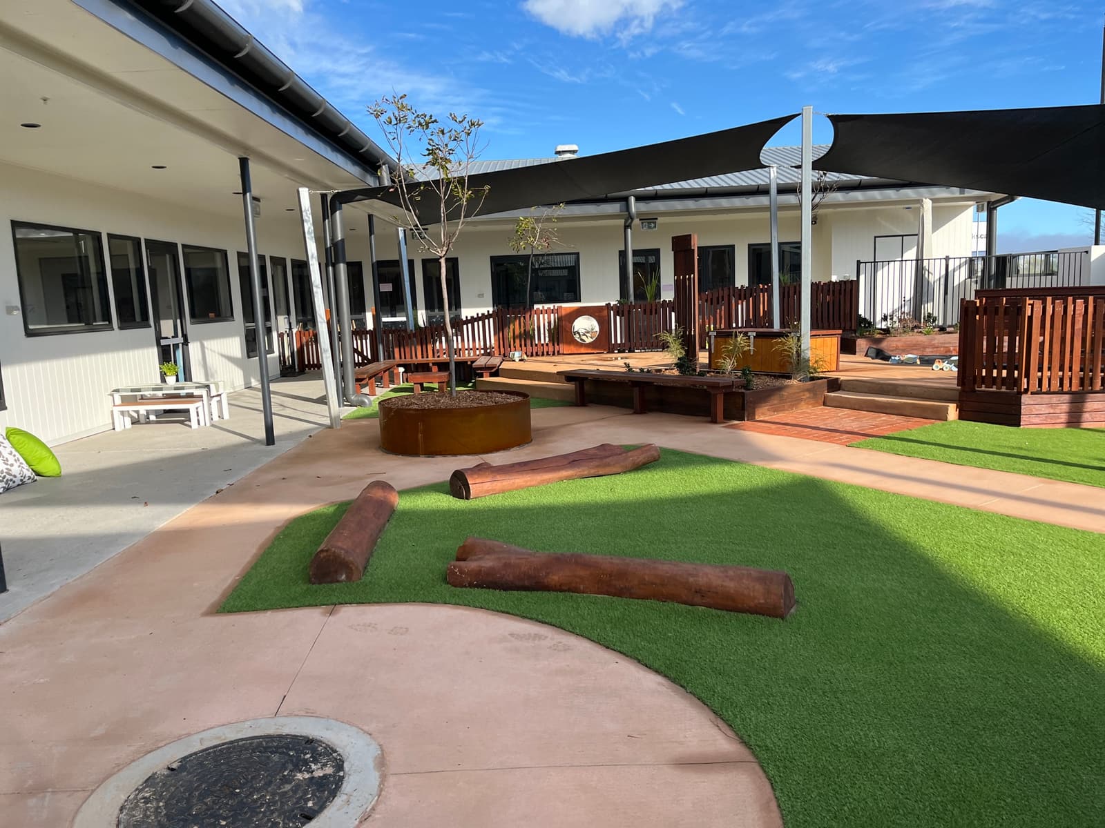 Courtyard in Toowoomba with log balance beams on synthetic turf, coloured concrete paths, timber decking with picket fencing and black shade sails