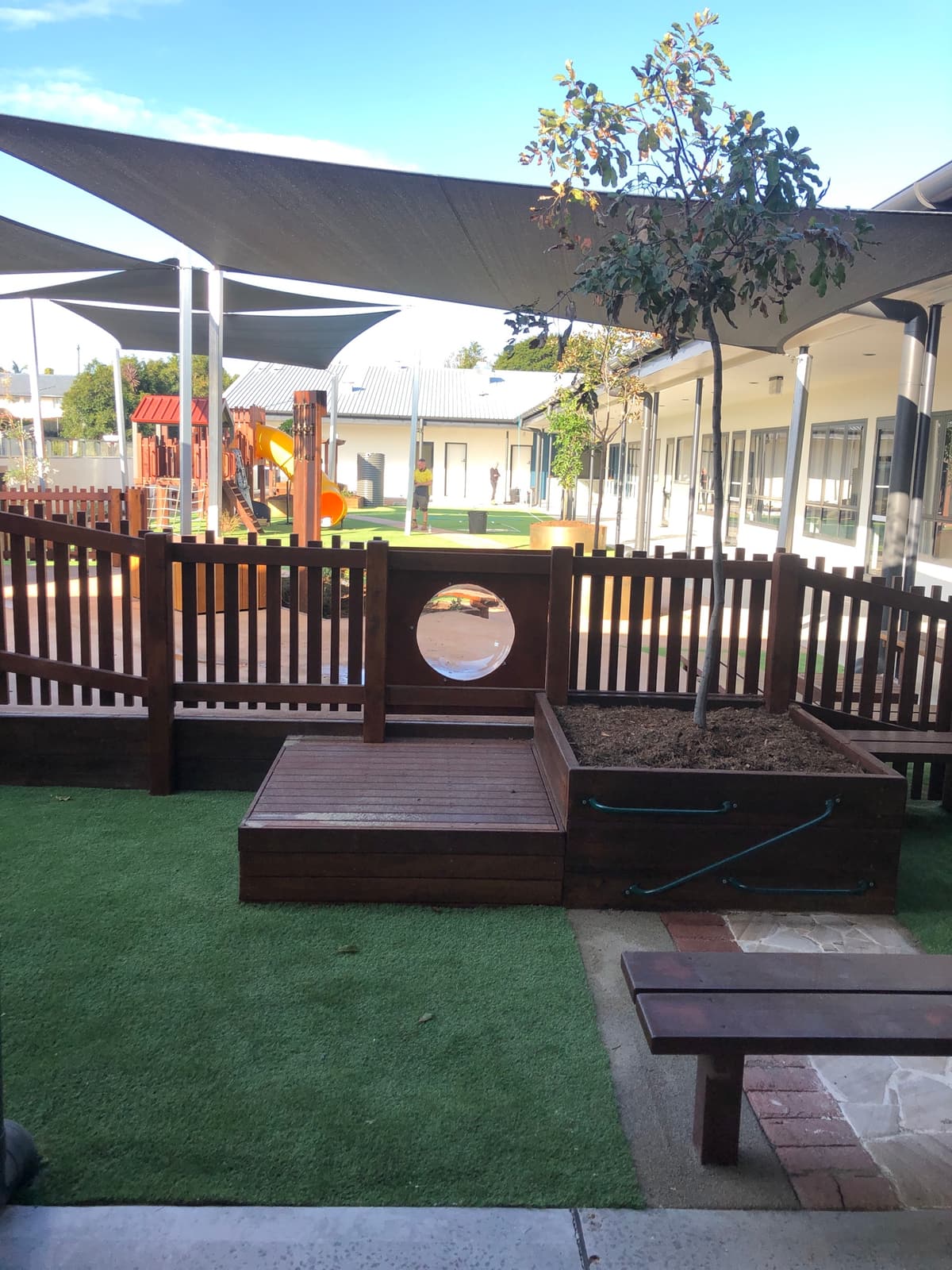 Raised timber deck and planter box with a porthole window in a timber picket fence, on synthetic turf, with a play fort and shade sails in the background in Toowoomba