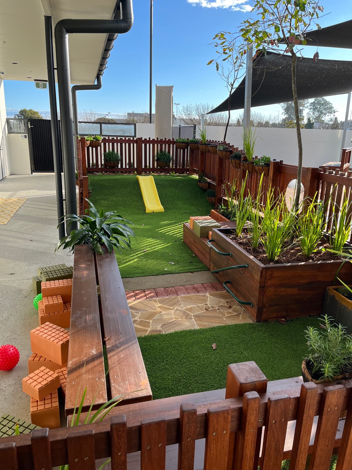Narrow side yard in Toowoomba with synthetic turf, a small yellow slide on a turf mound, timber planter boxes, a timber bench and sandstone crazy paving