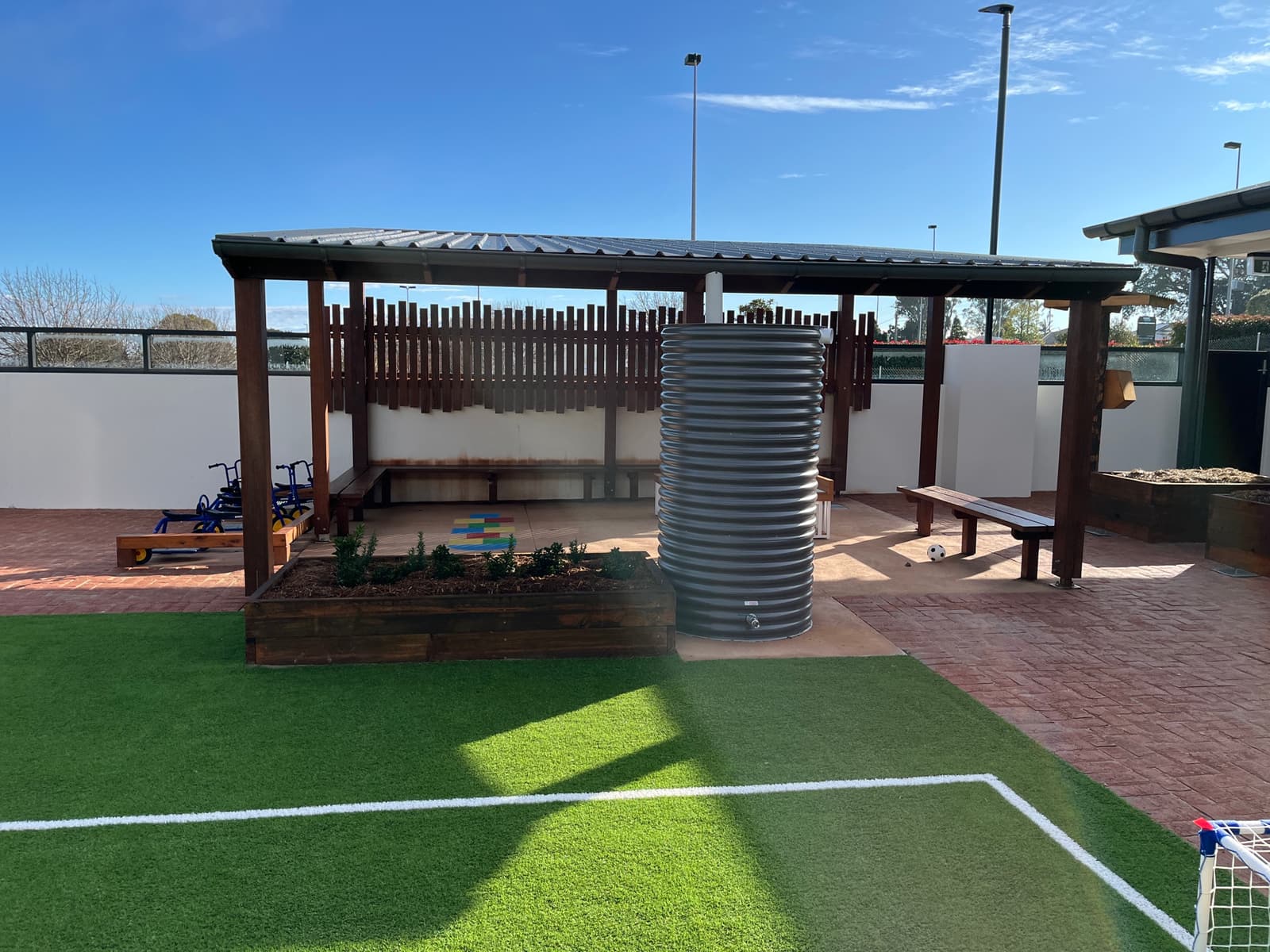 Timber-framed shelter with a metal roof, bench seating, a raised planter and a rainwater tank beside line-marked synthetic turf in Toowoomba